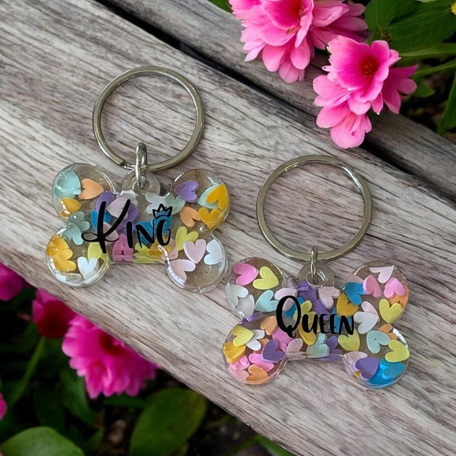 Personalised bone-shaped dog tags with colorful heart confetti, displayed on wood near pink flowers.