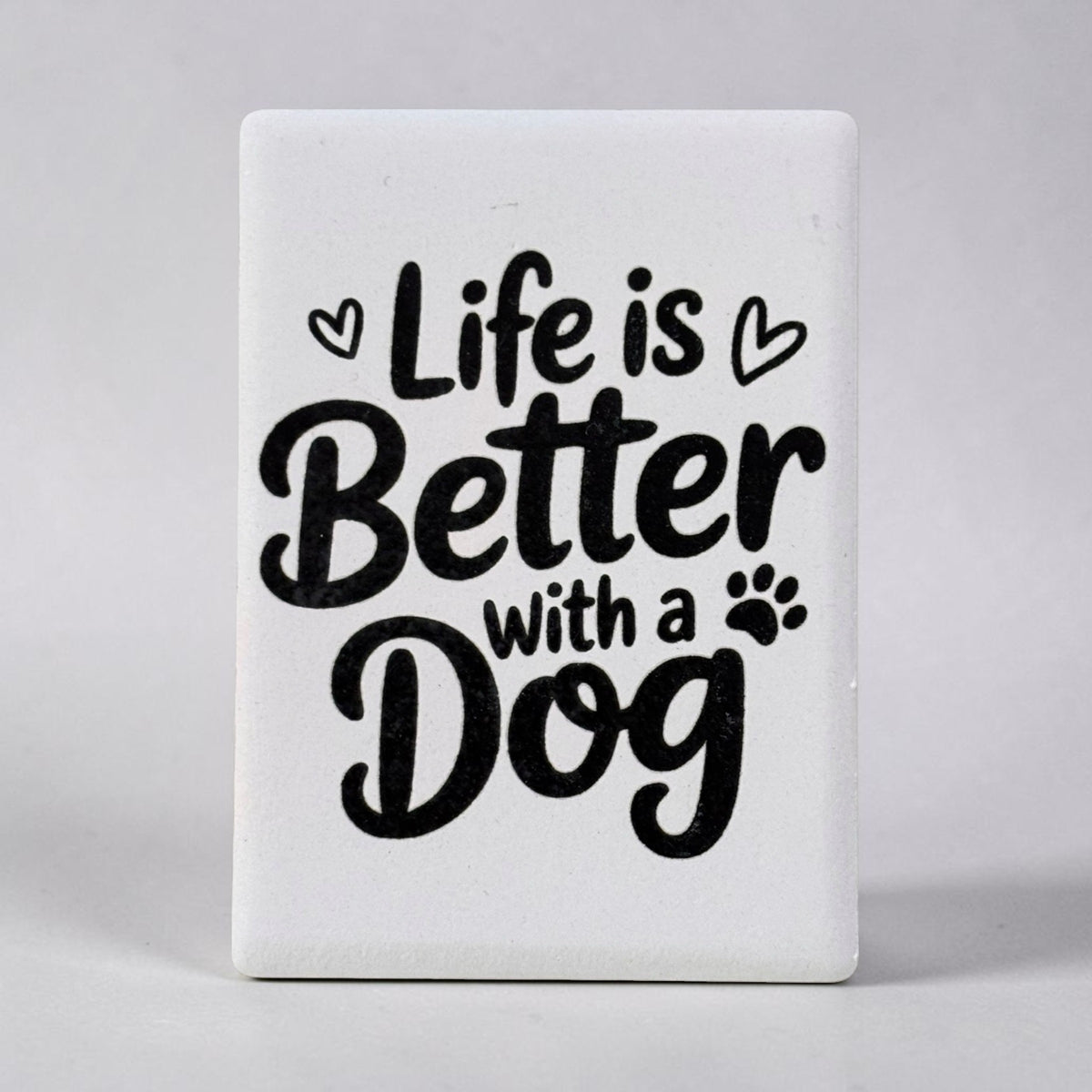 A white rectangular decorative magnet with rounded corners featuring the phrase 'Life is Better with a Dog' in black script and serif lettering, accented with small heart and paw print icons, shown against a neutral gray background.