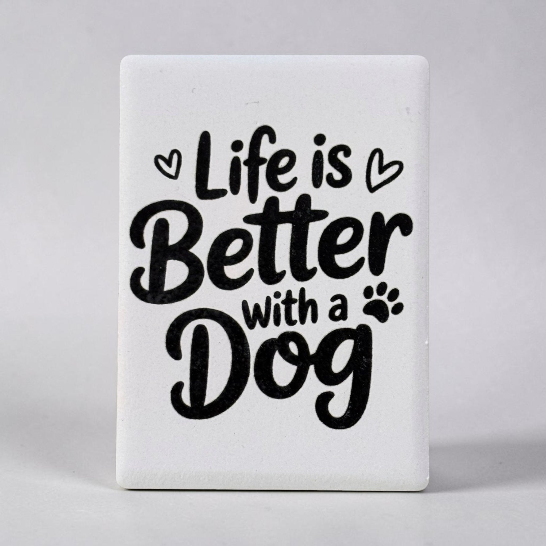 A white rectangular decorative magnet with rounded corners featuring the phrase 'Life is Better with a Dog' in black script and serif lettering, accented with small heart and paw print icons, shown against a neutral gray background.